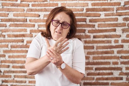 Téléchargez les photos : Femme âgée avec des lunettes debout sur le mur de briques souffrant de douleur aux mains et aux doigts, inflammation de l'arthrite - en image libre de droit