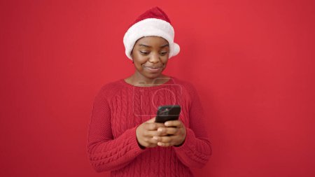 Téléchargez les photos : Femme afro-américaine portant un chapeau de Noël à l'aide d'un smartphone sur fond rouge isolé - en image libre de droit