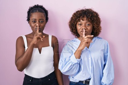 Téléchargez les photos : Deux femmes africaines debout sur fond rose demandant de se taire avec le doigt sur les lèvres. silence et concept secret. - en image libre de droit