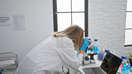 Photo for Attractive, focused blonde young woman scientist engrossed in her medical research using a microscope in lab, a portrait of determination working on scientific analysis - Royalty Free Image