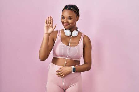 Téléchargez les photos : Femme afro-américaine avec des tresses portant des vêtements de sport et des écouteurs renonçant à dire bonjour heureux et souriant, geste de bienvenue amical - en image libre de droit