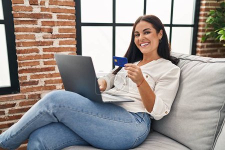 Foto de Joven mujer hispana hermosa usando portátil y tarjeta de crédito sentado en el sofá en casa - Imagen libre de derechos