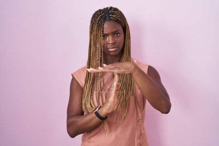Foto de Mujer afroamericana con cabello trenzado de pie sobre fondo rosa haciendo un gesto de tiempo fuera con las manos, rostro frustrado y serio - Imagen libre de derechos