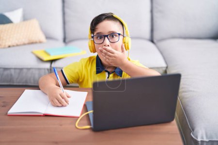 Foto de Joven niño hispano haciendo la tarea con el ordenador portátil en casa cubriendo la boca con la mano, sorprendido y con miedo a equivocarse. expresión sorprendida - Imagen libre de derechos