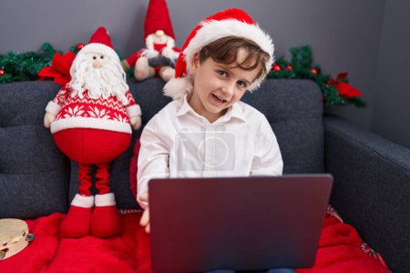 Photo for Adorable hispanic boy using laptop sitting on sofa by christmas decoration at home - Royalty Free Image