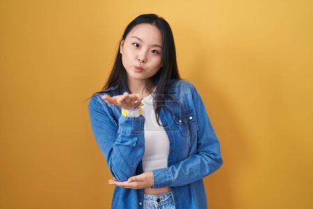 Téléchargez les photos : Jeune femme asiatique debout sur fond jaune regardant la caméra souffler un baiser avec la main sur l'air étant belle et sexy. expression d'amour. - en image libre de droit