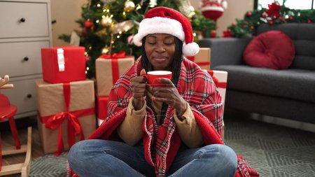 Photo for African woman drinking coffee sitting by christmas tree at home - Royalty Free Image