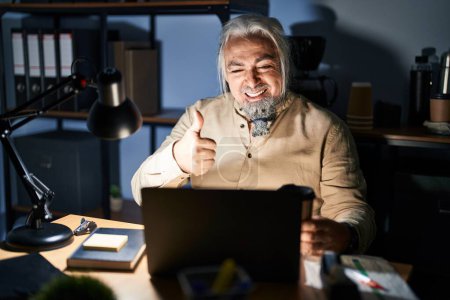 Téléchargez les photos : Homme d'âge moyen avec les cheveux gris qui travaillent au bureau la nuit en faisant des pouces heureux geste avec la main. approuvant l'expression regardant la caméra montrant le succès. - en image libre de droit