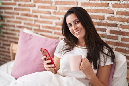 Téléchargez les photos : Jeune belle femme hispanique utilisant smartphone boire du café dans la chambre - en image libre de droit