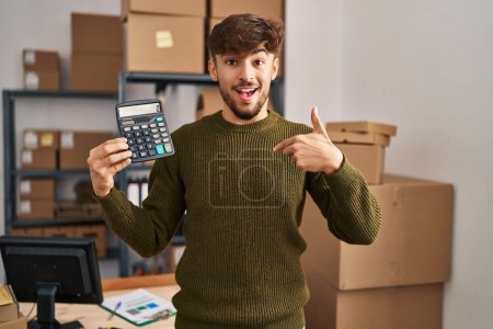 Téléchargez les photos : Arabe homme avec barbe travaillant à la petite entreprise commerce électronique tenant calculatrice pointant du doigt un sourire heureux et fier - en image libre de droit