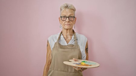 Foto de Artista madura de cabello gris, con pincel y paleta, pintando con seria precisión sobre caballete portátil en una relajada clase de arte aislada de paredes rosadas. - Imagen libre de derechos