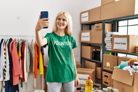 Téléchargez les photos : Jeune femme blonde bénévole faire selfie par smartphone souriant au centre de charité - en image libre de droit