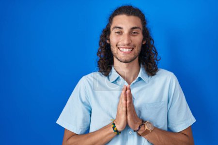 Téléchargez les photos : Jeune homme hispanique debout sur fond bleu priant avec les mains ensemble demandant pardon souriant confiant. - en image libre de droit