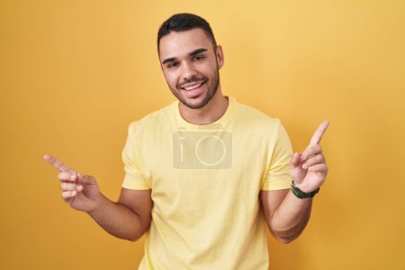 Téléchargez les photos : Jeune homme hispanique debout sur fond jaune souriant confiant pointant avec les doigts vers différentes directions. espace de copie pour la publicité - en image libre de droit