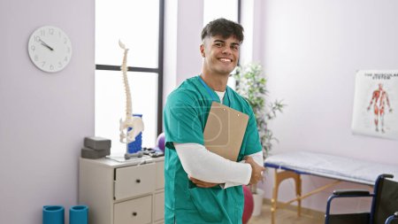 Téléchargez les photos : Un jeune homme hispanique confiant, un physiothérapeute rayonnant, affiche fièrement son document de traitement de réadaptation dans une clinique de réadaptation animée - en image libre de droit