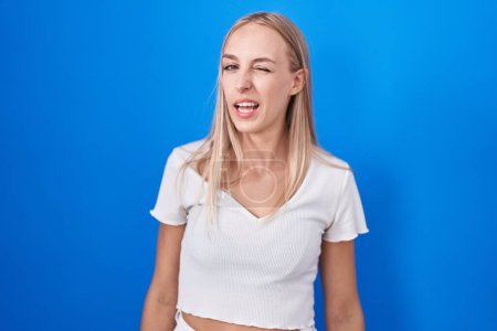 Foto de Young caucasian woman standing over blue background winking looking at the camera with sexy expression, cheerful and happy face. - Imagen libre de derechos