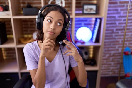 Photo for Young hispanic woman playing video games wearing headphones serious face thinking about question with hand on chin, thoughtful about confusing idea - Royalty Free Image