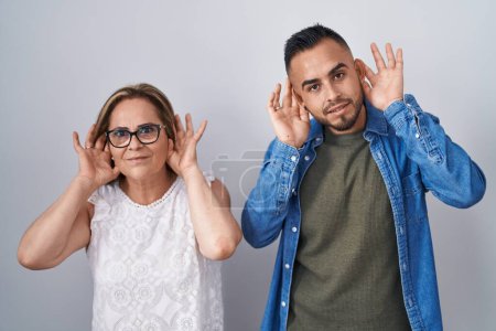 Téléchargez les photos : Mère hispanique et fils debout ensemble en essayant d'entendre les deux mains sur le geste de l'oreille, curieux de commérages. problème auditif, sourd - en image libre de droit