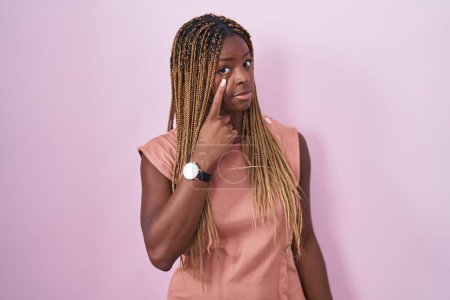 Téléchargez les photos : Femme afro-américaine avec les cheveux tressés debout sur fond rose pointant vers l'oeil vous regardant geste, expression suspecte - en image libre de droit
