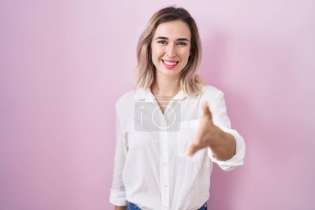 Foto de Joven hermosa mujer de pie sobre fondo rosa sonriendo amistoso ofreciendo apretón de manos como saludo y bienvenida. negocios exitosos. - Imagen libre de derechos