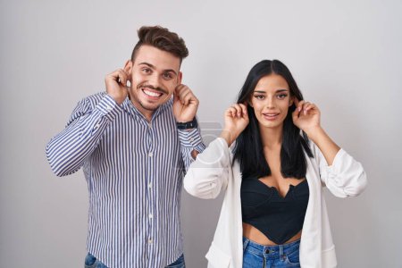 Photo for Young hispanic couple standing over white background smiling pulling ears with fingers, funny gesture. audition problem - Royalty Free Image