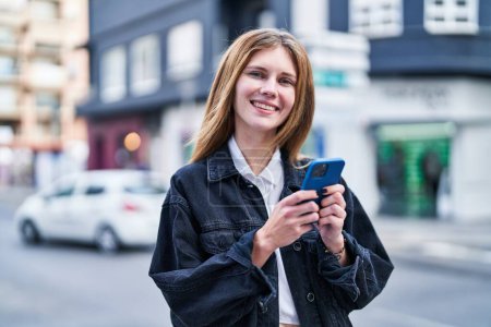 Foto de Mujer rubia joven usando teléfono inteligente sonriendo en la calle - Imagen libre de derechos
