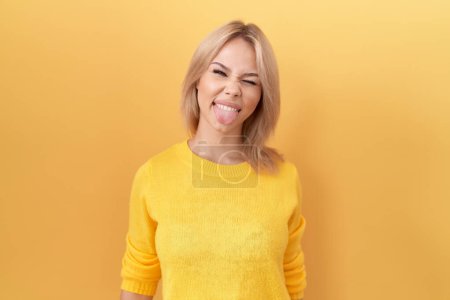 Téléchargez les photos : Jeune femme caucasienne portant un pull jaune qui sort la langue heureuse avec une expression drôle. concept d'émotion. - en image libre de droit