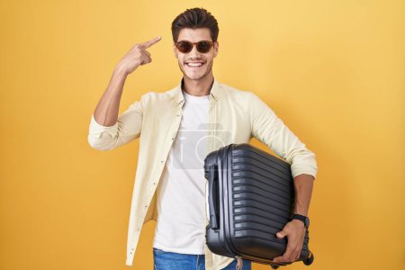 Téléchargez les photos : Jeune homme hispanique tenant une valise allant en vacances d'été souriant pointant vers la tête avec un doigt, grande idée ou pensée, bon souvenir - en image libre de droit
