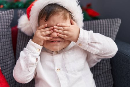 Photo for Adorable hispanic toddler wearing christmas hat covering eyes with hands at home - Royalty Free Image