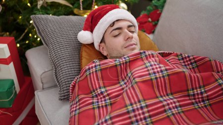 Foto de Joven hombre hispano durmiendo tumbado en el sofá por el árbol de Navidad en casa - Imagen libre de derechos