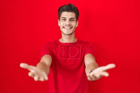 Téléchargez les photos : Jeune homme hispanique debout sur fond rouge souriant joyeuse offrant des mains donnant assistance et acceptation. - en image libre de droit