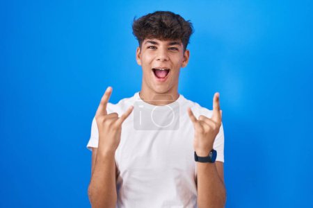 Photo for Hispanic teenager standing over blue background shouting with crazy expression doing rock symbol with hands up. music star. heavy music concept. - Royalty Free Image
