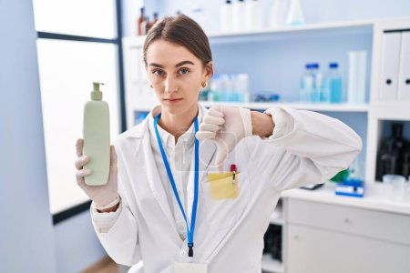 Photo for Young caucasian woman working at scientist laboratory holding body lotion with angry face, negative sign showing dislike with thumbs down, rejection concept - Royalty Free Image