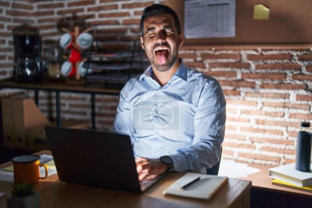Téléchargez les photos : Homme hispanique avec barbe travaillant au bureau la nuit langue collante heureux avec une expression drôle. concept d'émotion. - en image libre de droit