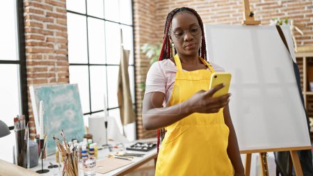 Téléchargez les photos : Belle artiste afro-américaine peint son chef-d'œuvre en toile, debout dans un tablier avec son smartphone dans un centre d'apprentissage atelier d'art - en image libre de droit