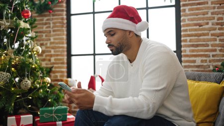 Foto de Hombre afroamericano usando teléfono inteligente celebrando la Navidad buscando molesto en casa - Imagen libre de derechos