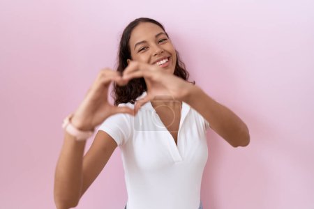 Téléchargez les photos : Jeune femme hispanique portant un t-shirt blanc décontracté souriant en amour faisant forme de symbole de coeur avec les mains. concept romantique. - en image libre de droit