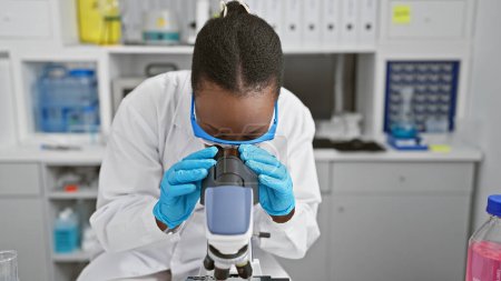 Foto de Científica afroamericana enfocada absorta en la investigación científica, en el análisis del microscopio en un laboratorio bullicioso; sus gafas simbolizan una búsqueda de descubrimiento médico. - Imagen libre de derechos