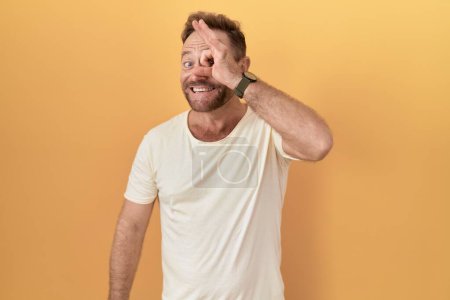 Photo for Middle age man with beard standing over yellow background doing ok gesture with hand smiling, eye looking through fingers with happy face. - Royalty Free Image