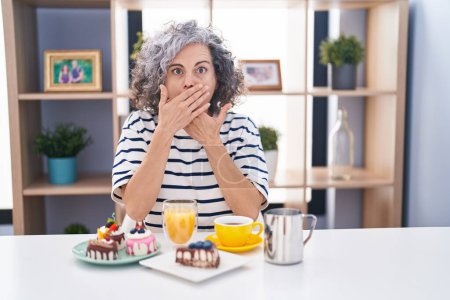 Téléchargez les photos : Femme d'âge moyen avec les cheveux gris manger des pâtisseries et boire du café pour le petit déjeuner choqué couvrant la bouche avec les mains pour erreur. concept secret. - en image libre de droit
