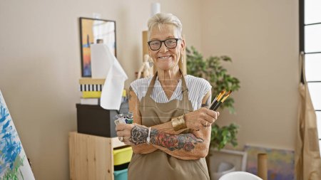 Téléchargez les photos : Femme âgée aux cheveux gris souriante debout avec un geste de bras croisés détendu, jouissant de son passe-temps au milieu de la toile et des pinceaux dans son atelier d'art. - en image libre de droit