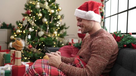 Foto de Joven hombre caucásico usando teléfono inteligente sentado en el sofá por el árbol de Navidad en casa - Imagen libre de derechos