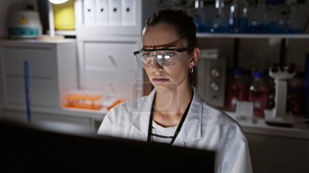 Foto de Joven hermosa mujer hispana científica usando anteojos usando computadora sonriendo en el laboratorio - Imagen libre de derechos