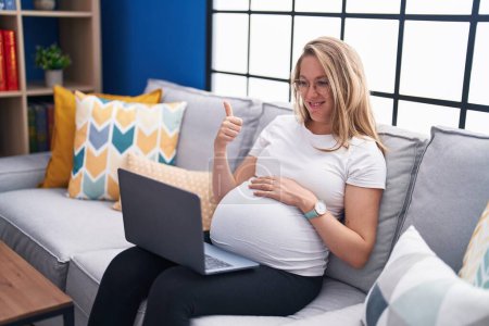 Foto de Mujer embarazada joven sentada en el sofá en casa usando el ordenador portátil sonriendo feliz y positivo, pulgar hacia arriba haciendo excelente y signo de aprobación - Imagen libre de derechos