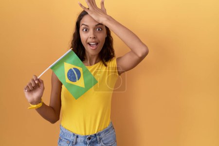 Foto de Mujer hispana joven sosteniendo bandera de Brasil sorprendida con la mano en la cabeza por error, recuerde el error. olvidado, mal concepto de memoria. - Imagen libre de derechos