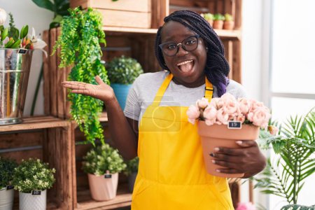 Téléchargez les photos : Jeune femme africaine travaillant au magasin de fleuriste tenant usine célébrant la réalisation avec sourire heureux et expression gagnante avec la main levée - en image libre de droit