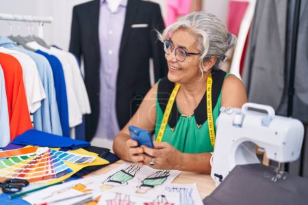 Photo for Middle age grey-haired woman tailor smiling confident using smartphone at tailor shop - Royalty Free Image
