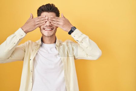 Téléchargez les photos : Jeune homme hispanique debout sur fond jaune couvrant les yeux avec les mains souriantes gaies et drôles. concept aveugle. - en image libre de droit