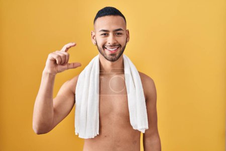 Téléchargez les photos : Jeune homme hispanique debout torse nu avec serviette geste souriant et confiant avec la main faisant signe de petite taille avec les doigts regardant et la caméra. concept de mesure. - en image libre de droit
