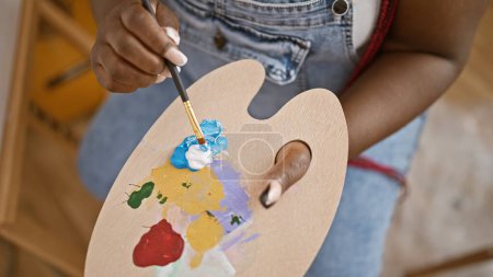 Foto de En una clase de arte interior conmovedora, una artista afroamericana mezcla hábilmente el color en su paleta, sus manos bailando sobre el lienzo en su estudio con pinceles - Imagen libre de derechos